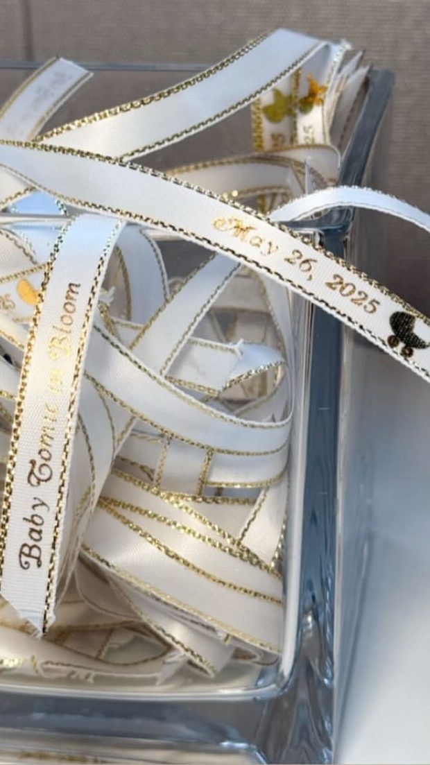 Loops of white satin ribbon printed in gold for the baby shower, gathered in a clear box.