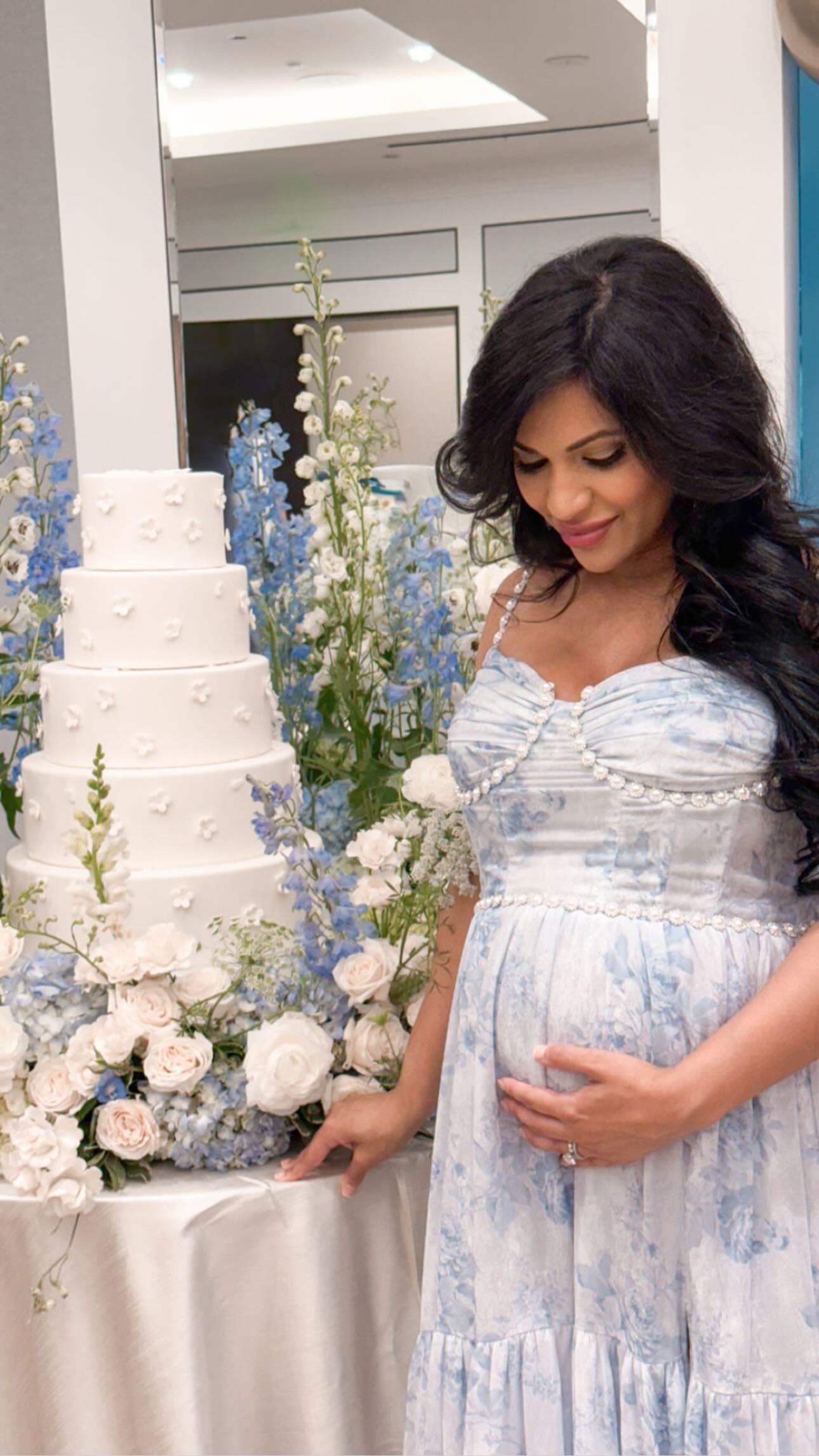 A mother-to-be in a blue and white floral gown stands beside a tall white tiered cake surrounded by blue delphinium and white roses.