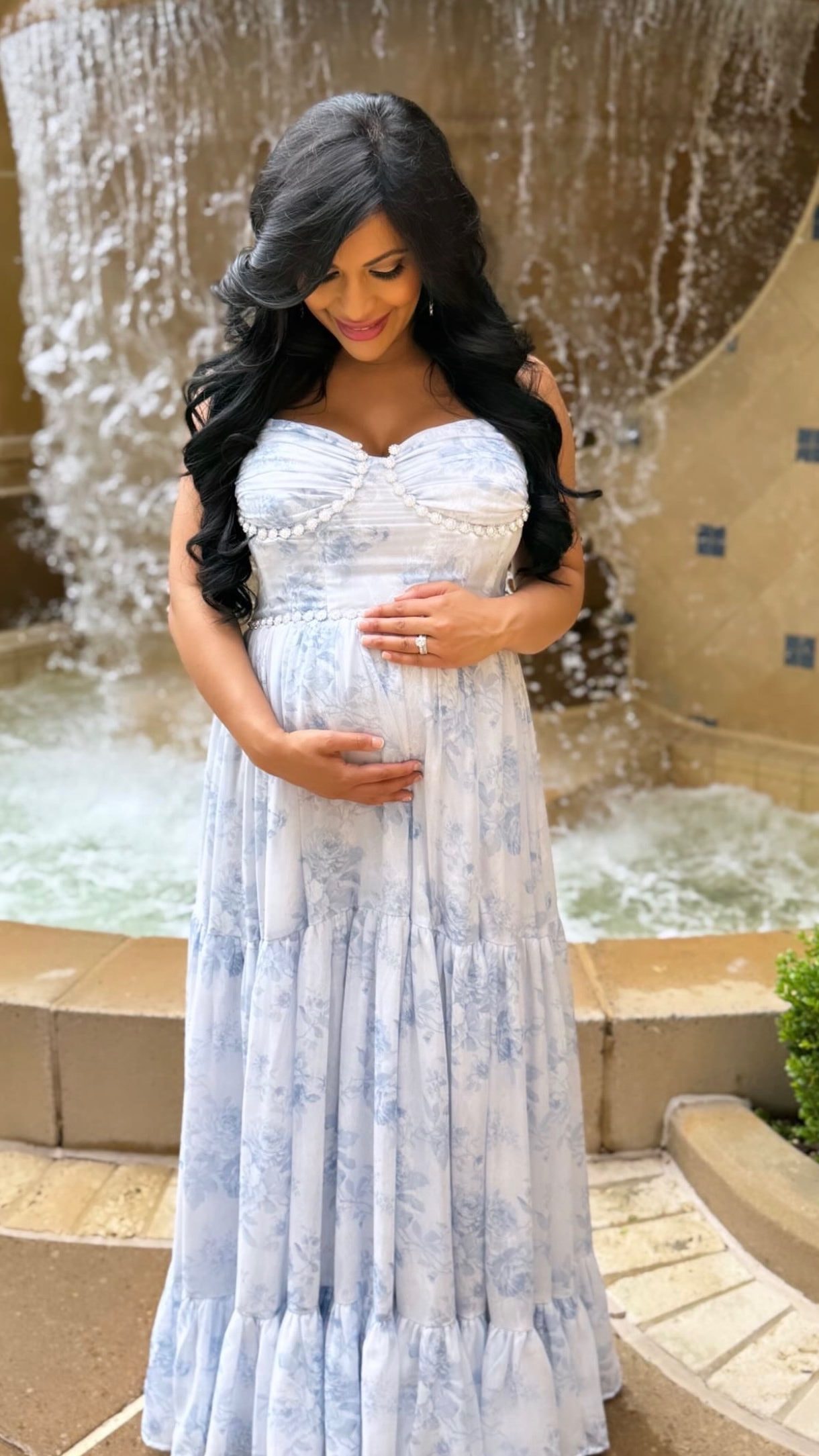 A mother-to-be in a long blue and white floral gown cradles her bump in front of a tiled courtyard fountain.