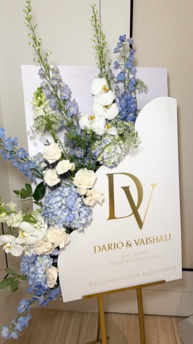 A white welcome sign with a gold monogram on a gold easel, framed by blue delphinium and white flowers.