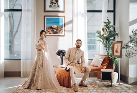 A bride in an ivory embroidered lehenga with a sheer dupatta stands beside a groom in a beige sherwani seated on a rust-colored ottoman in a bright, modern lounge with tall windows and framed art.