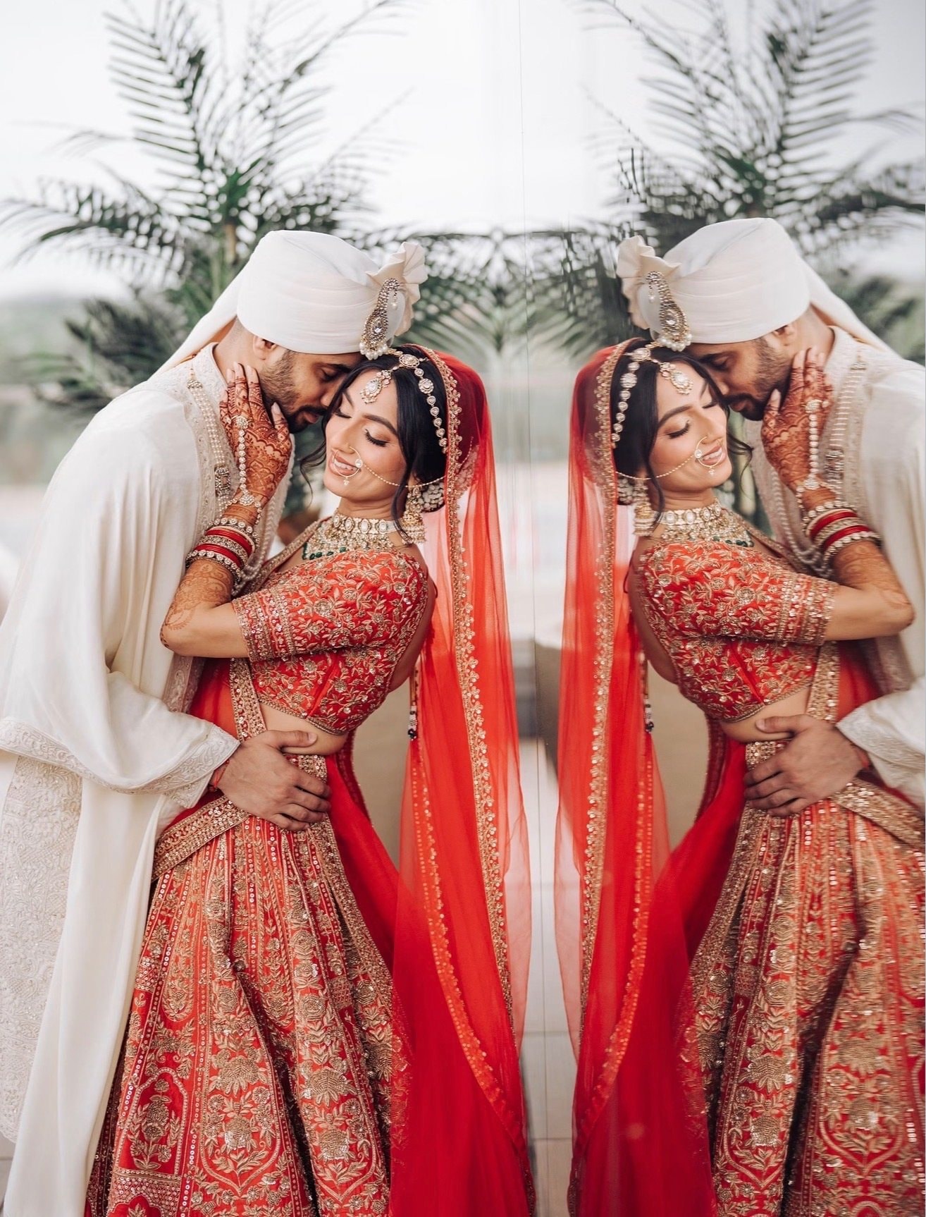 A mirrored image of a bride in a red embroidered lehenga and dupatta held by a groom in an ivory sherwani and pale pink turban, with palm fronds behind them.