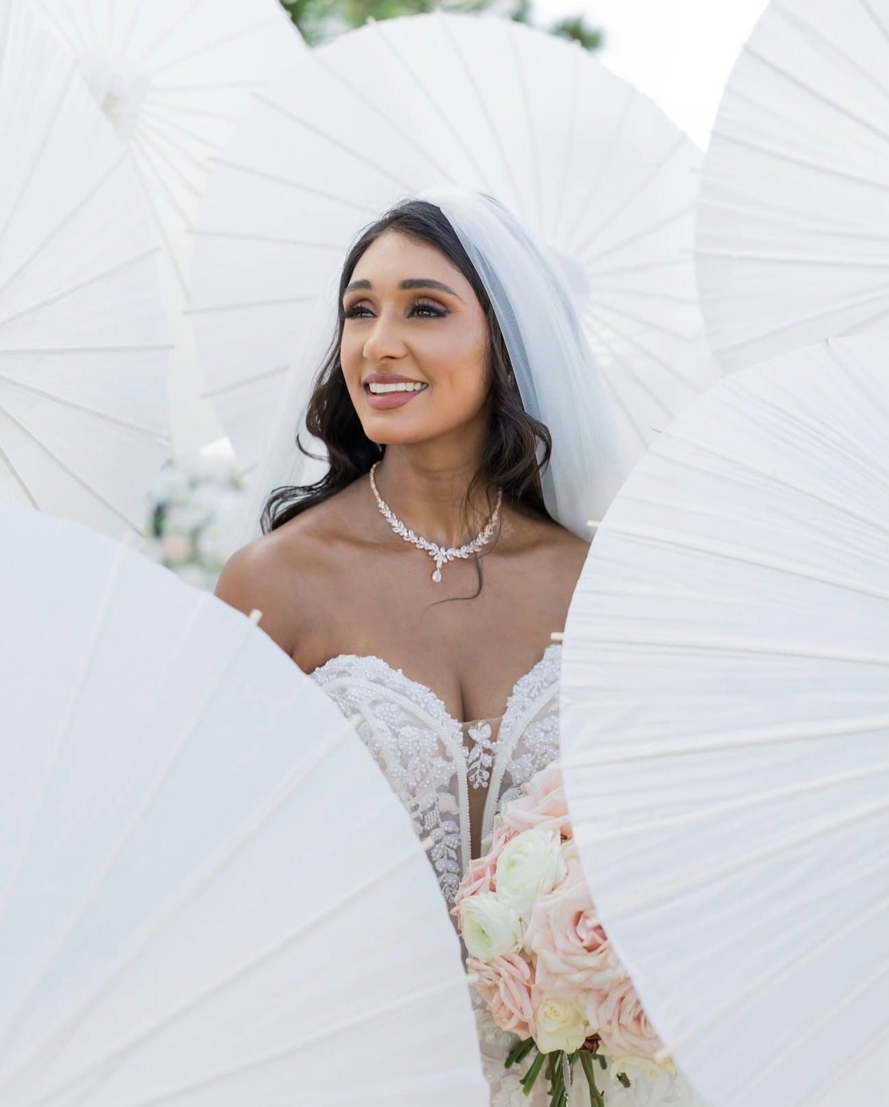 A smiling bride in a white lace gown and veil, holding a blush bouquet, framed by white paper parasols.