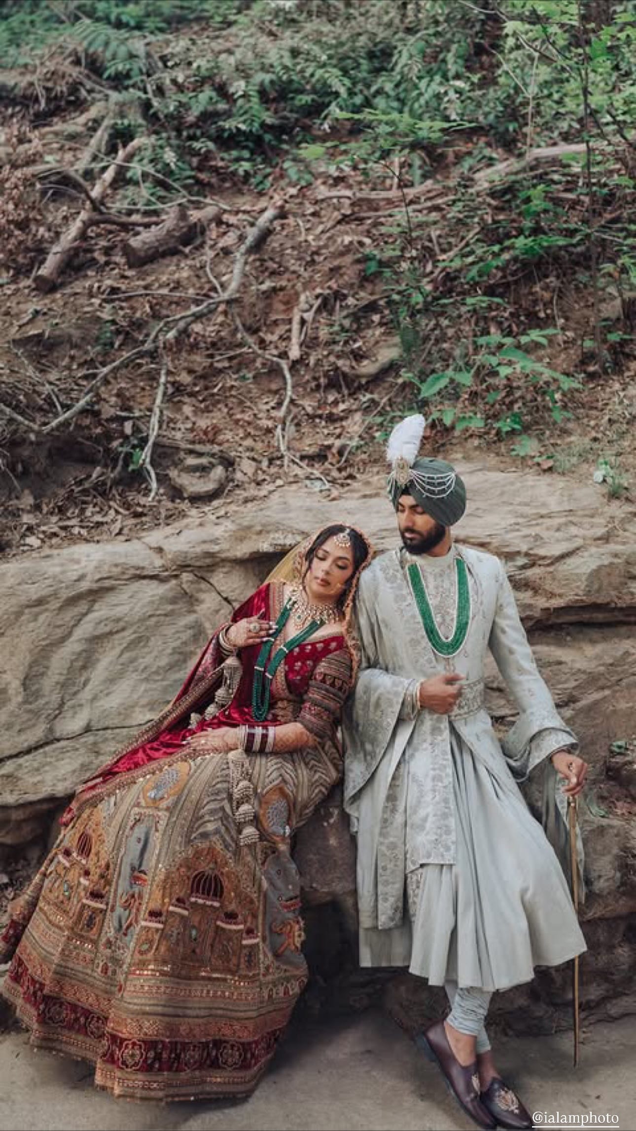 A bride in a red velvet dupatta and an embroidered lehenga rests her head on a groom in a pale grey sherwani, sage turban and emerald beads as they sit on a rock ledge in the woods.