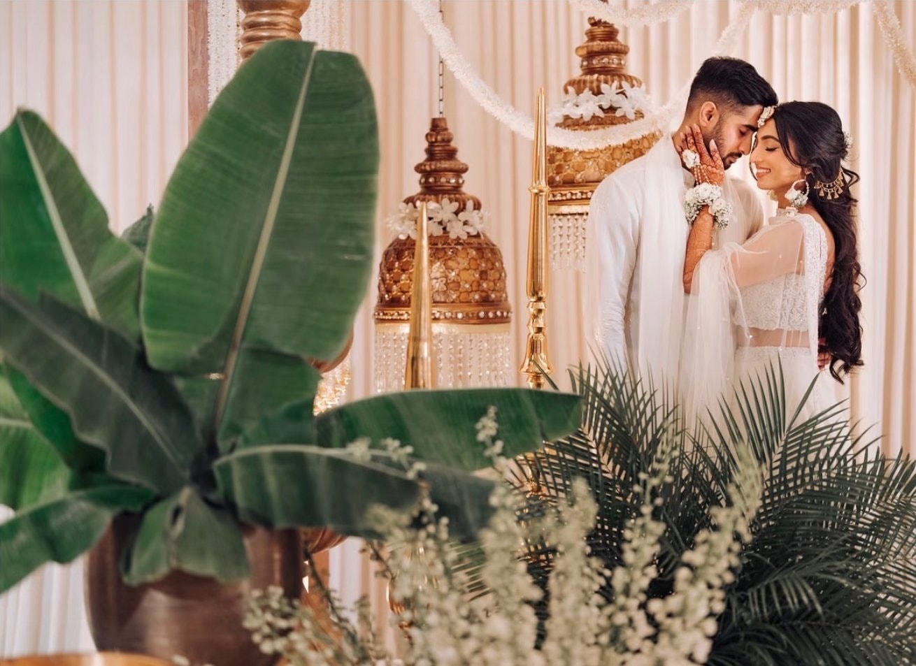 A groom in an ivory sherwani rests his forehead against his bride’s, she in a white embroidered outfit, beneath brass lanterns and between banana leaves and palms.