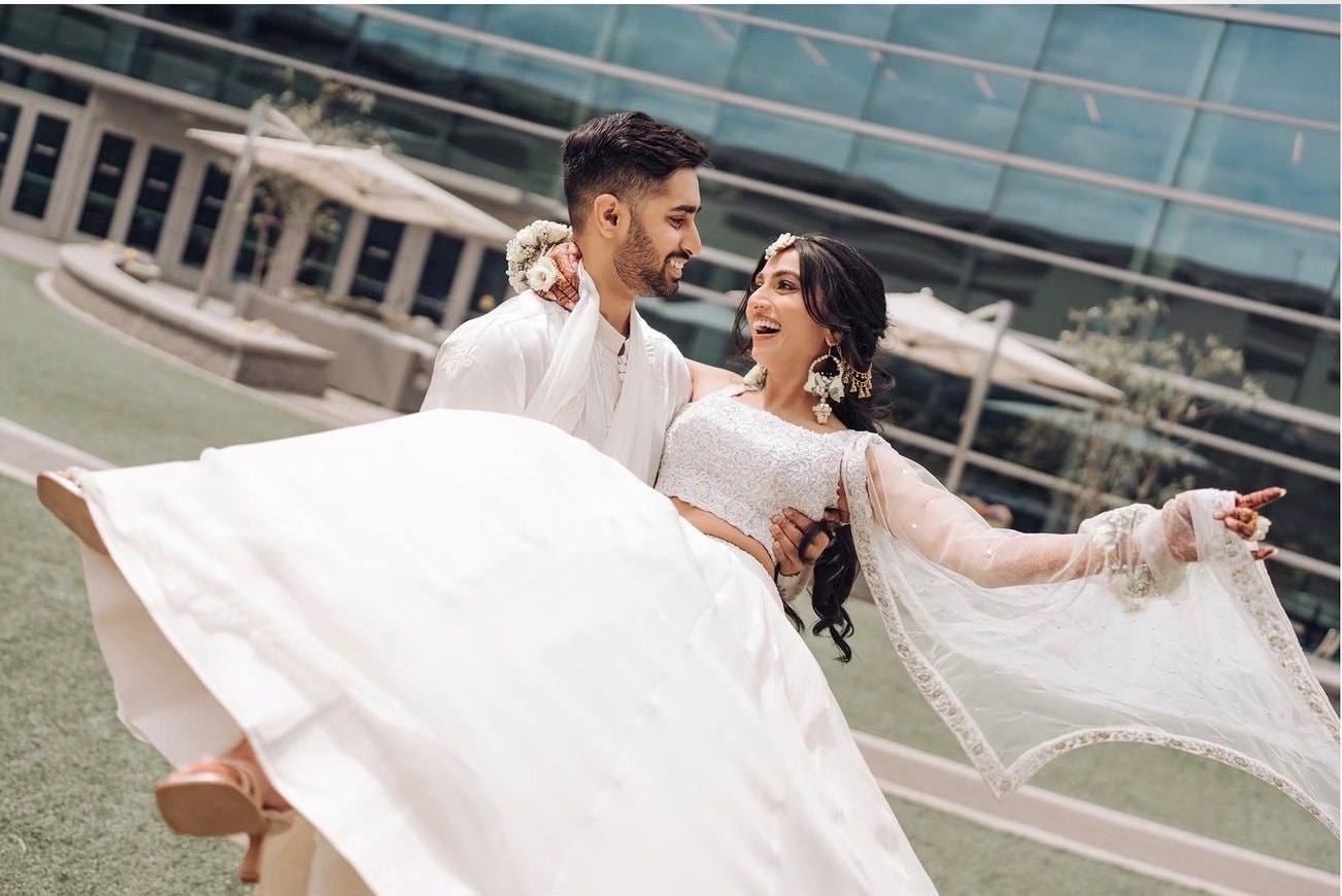 A groom in an ivory sherwani carries his laughing bride in a white lehenga across a lawn, her feet kicked up and one arm outstretched as her sheer dupatta flows.