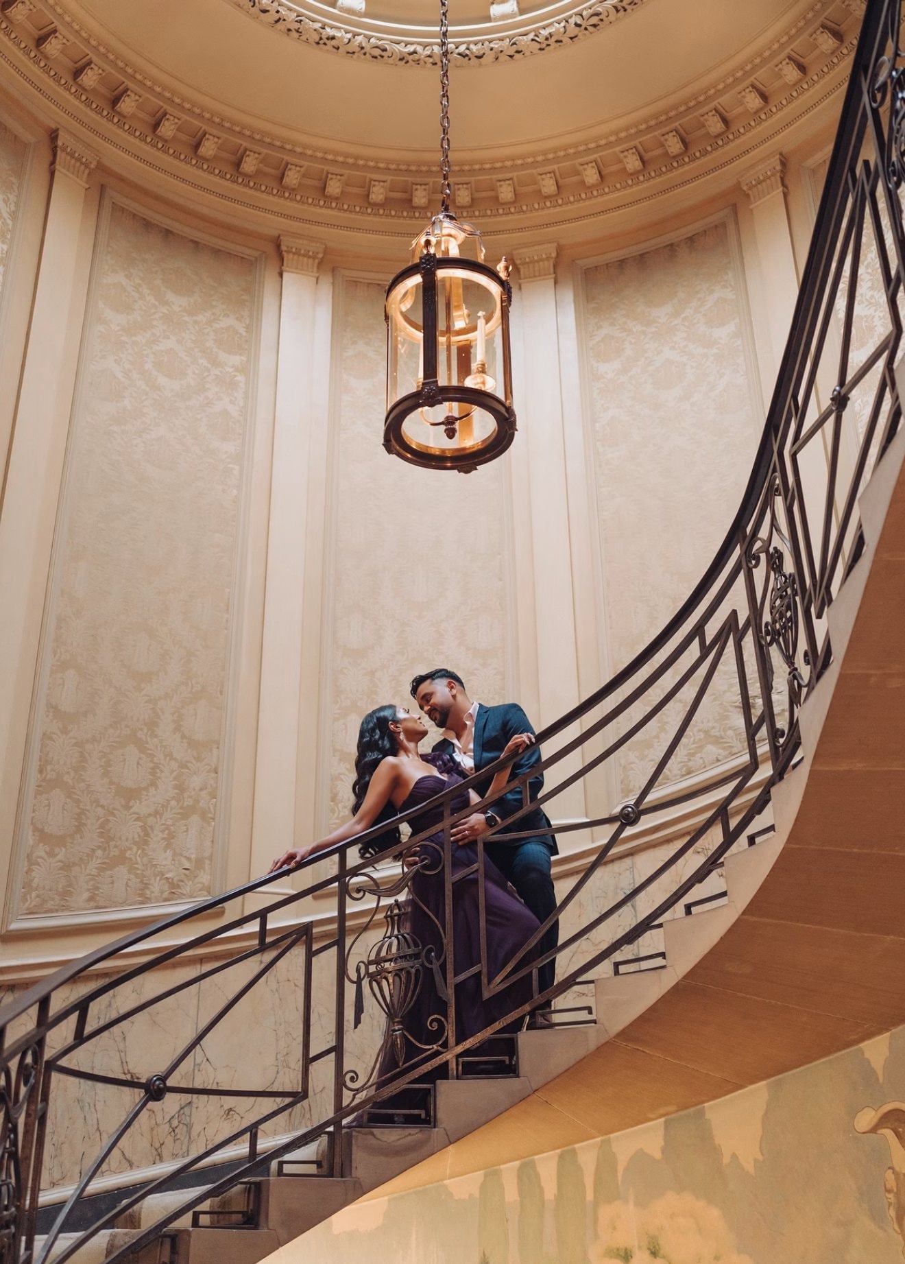 A couple shares a moment on a curving wrought-iron staircase beneath a domed ceiling and a hanging lantern, the woman in a long purple gown and the man in a navy suit.