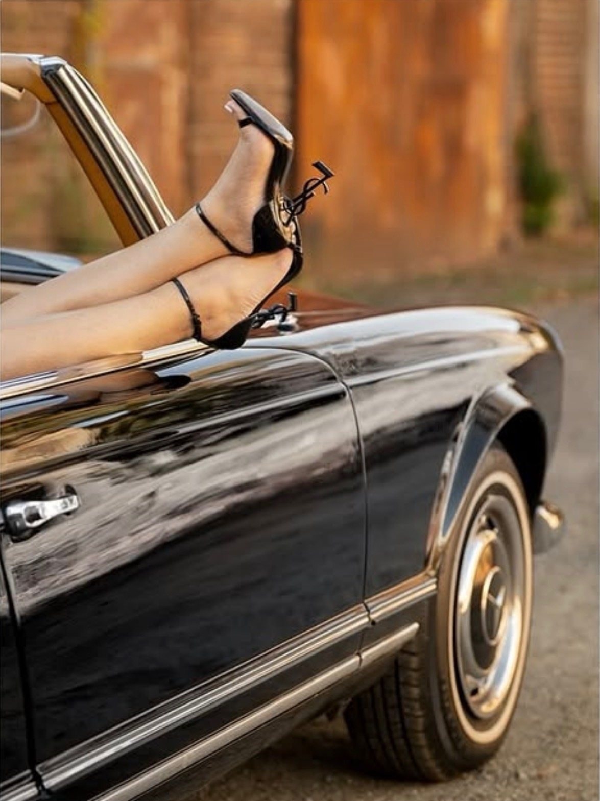 A woman’s legs in black strappy heels rest over the door of a black vintage convertible.