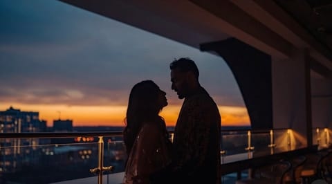 A couple in silhouette faces each other on a rooftop terrace at dusk, city lights and an orange and violet sky behind a glass railing.