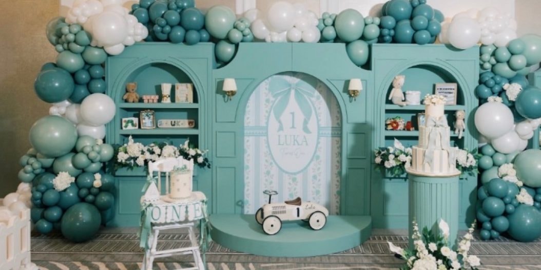 A teal first-birthday backdrop with arched shelves of toys, a white high chair with a ONE banner, a toy race car and a tiered cake, framed by balloon garlands in teal, sage and white.