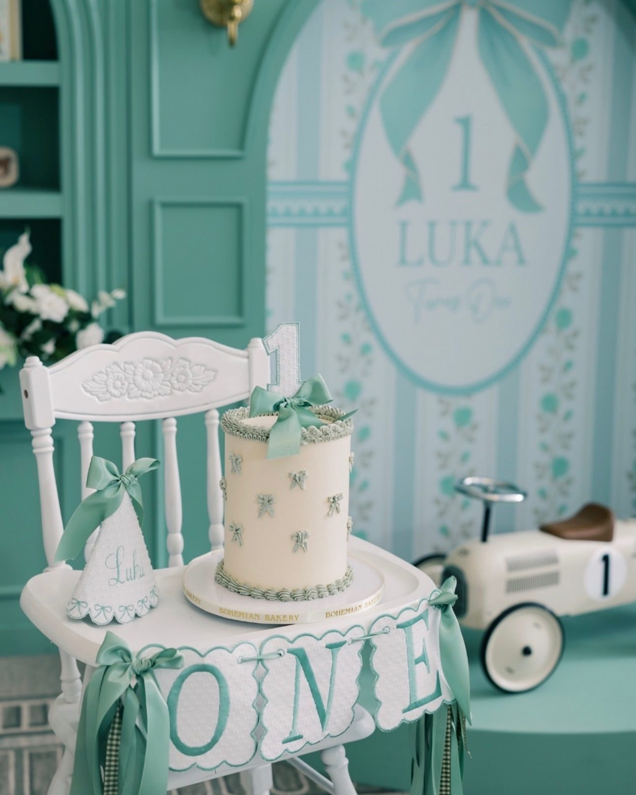A small white smash cake with sage bows on a white high chair hung with a ONE banner, beside a party hat and a toy race car.