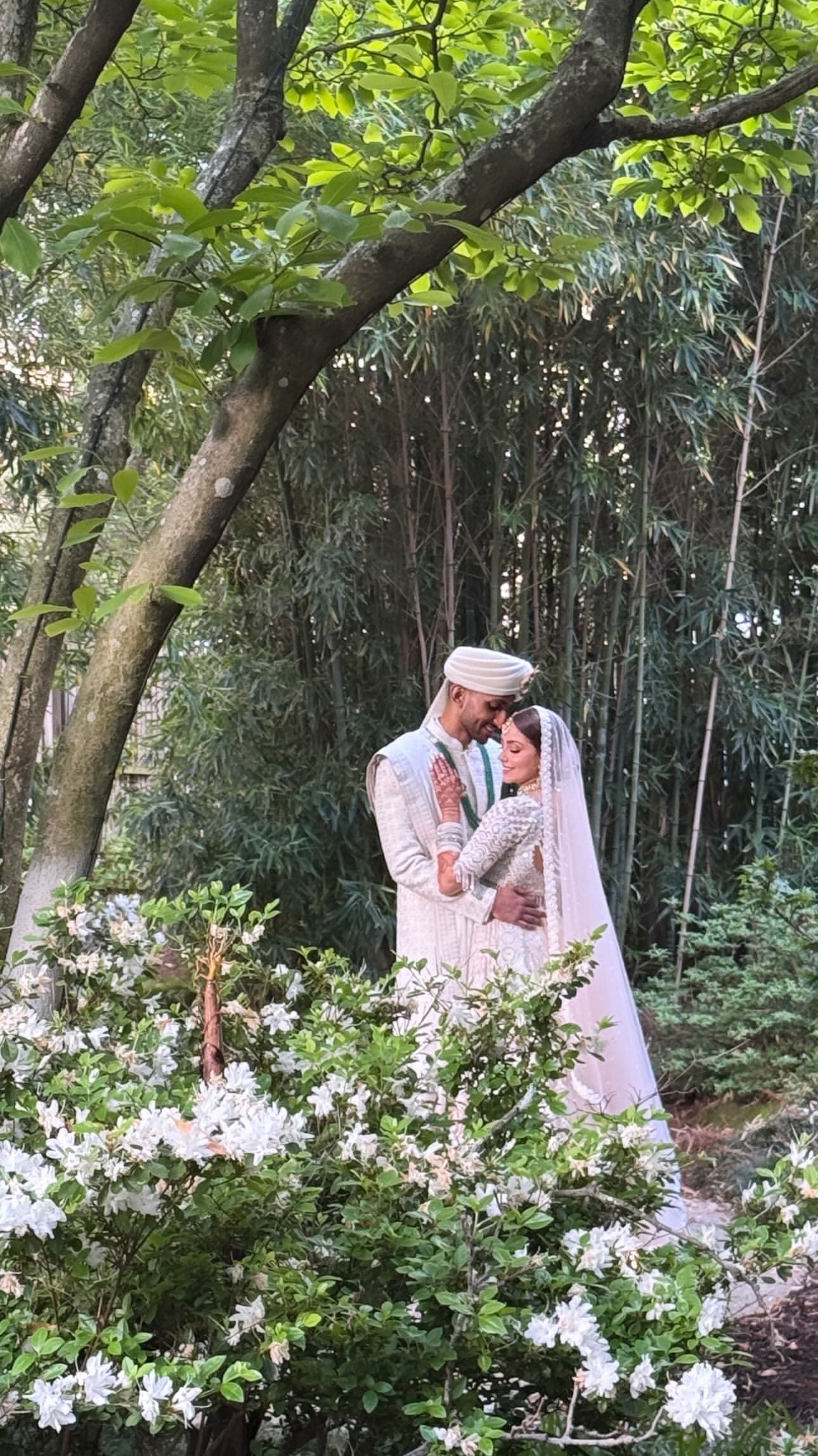 A groom in an ivory sherwani and cream turban holds a bride in a white embroidered bridal outfit and veil beneath a tree, with white azalea blossoms in the foreground and bamboo behind them.