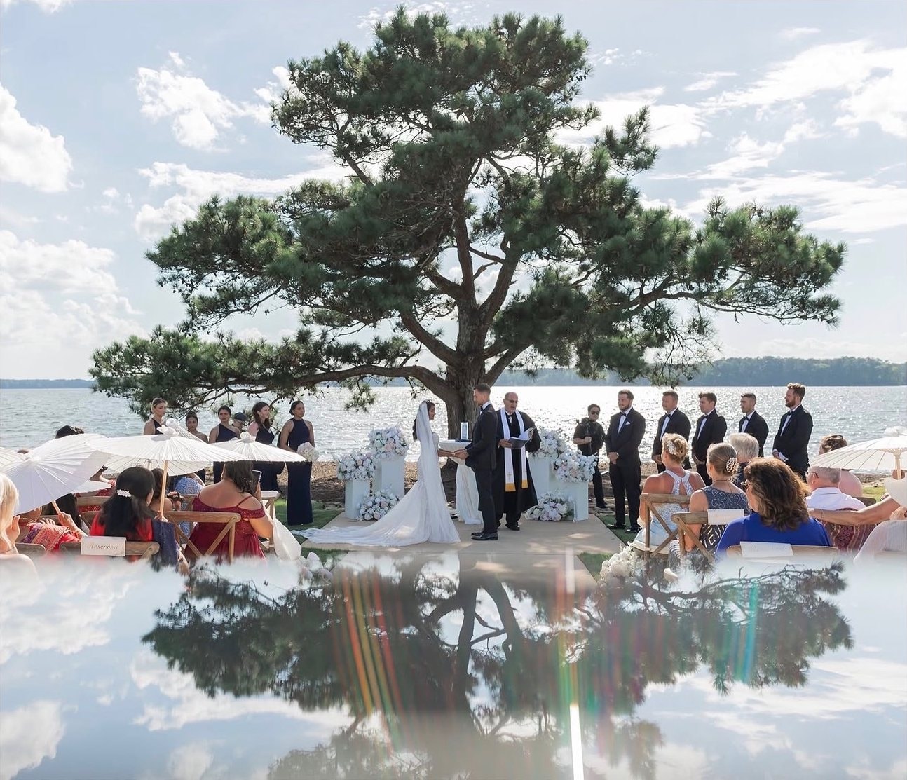 Guests watch a ceremony on a waterfront lawn beneath a large spreading tree, with the scene and sky reflected below.