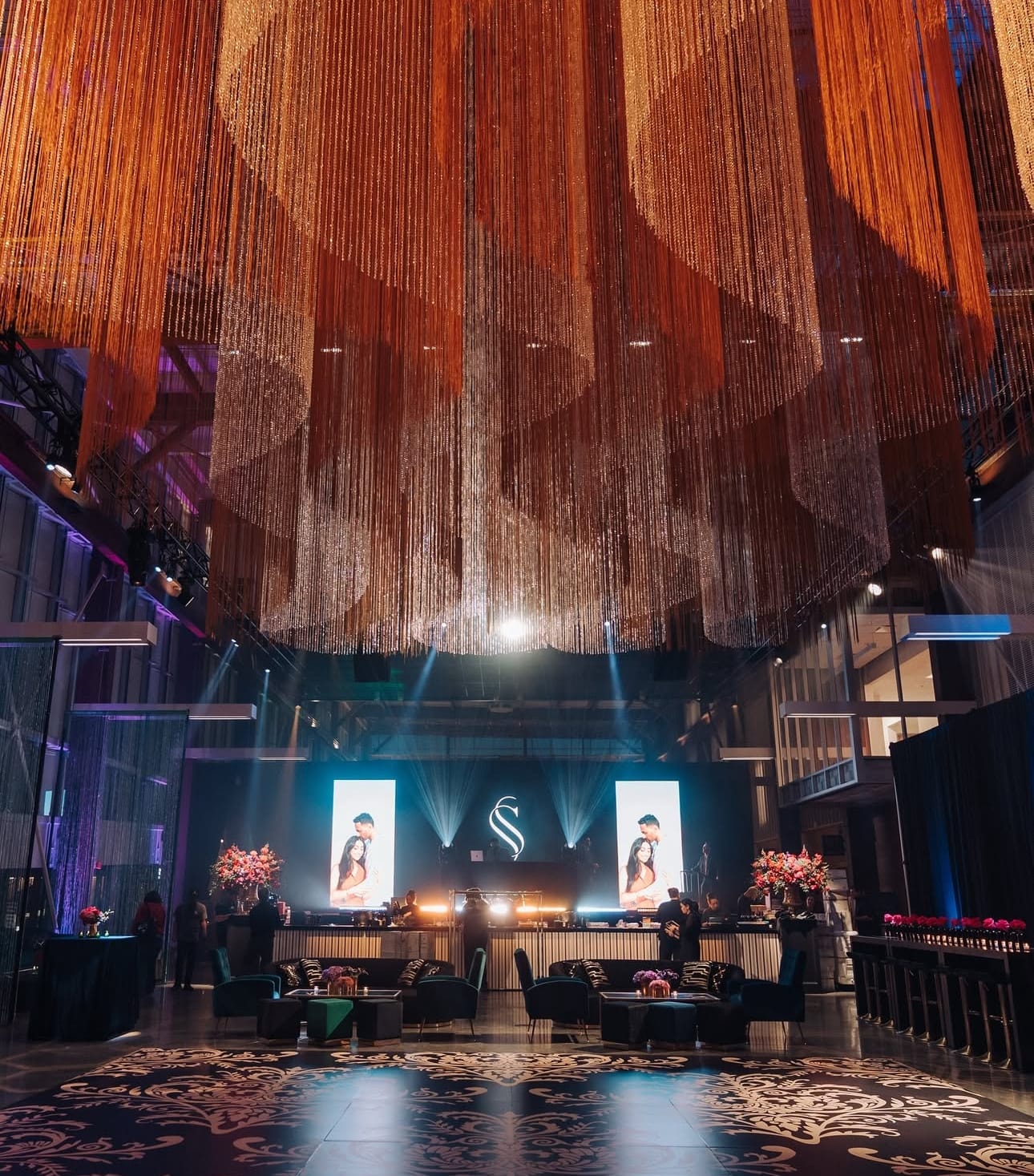 A reception hall with a vast copper-toned string installation hanging from the ceiling, a lit stage with a monogram and portrait screens, lounge seating, and a patterned dance floor.