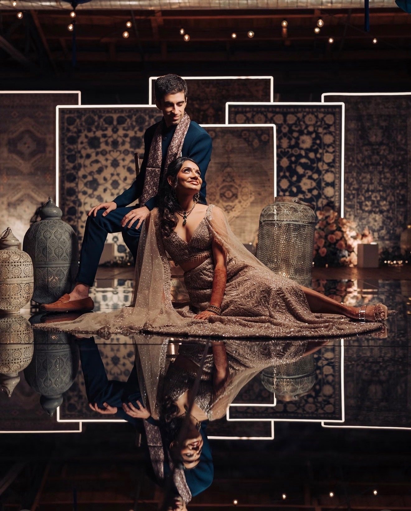 A couple in formal Indian wear poses on a mirrored floor in front of layered rugs framed by glowing light strips, the man in a navy sherwani and the woman in a beaded lehenga.