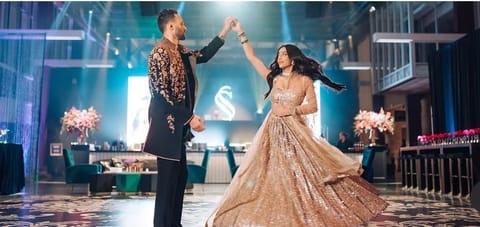 A groom in a black embroidered sherwani twirls a bride in a champagne sequined lehenga on a patterned dance floor beneath blue stage lighting and a monogram.