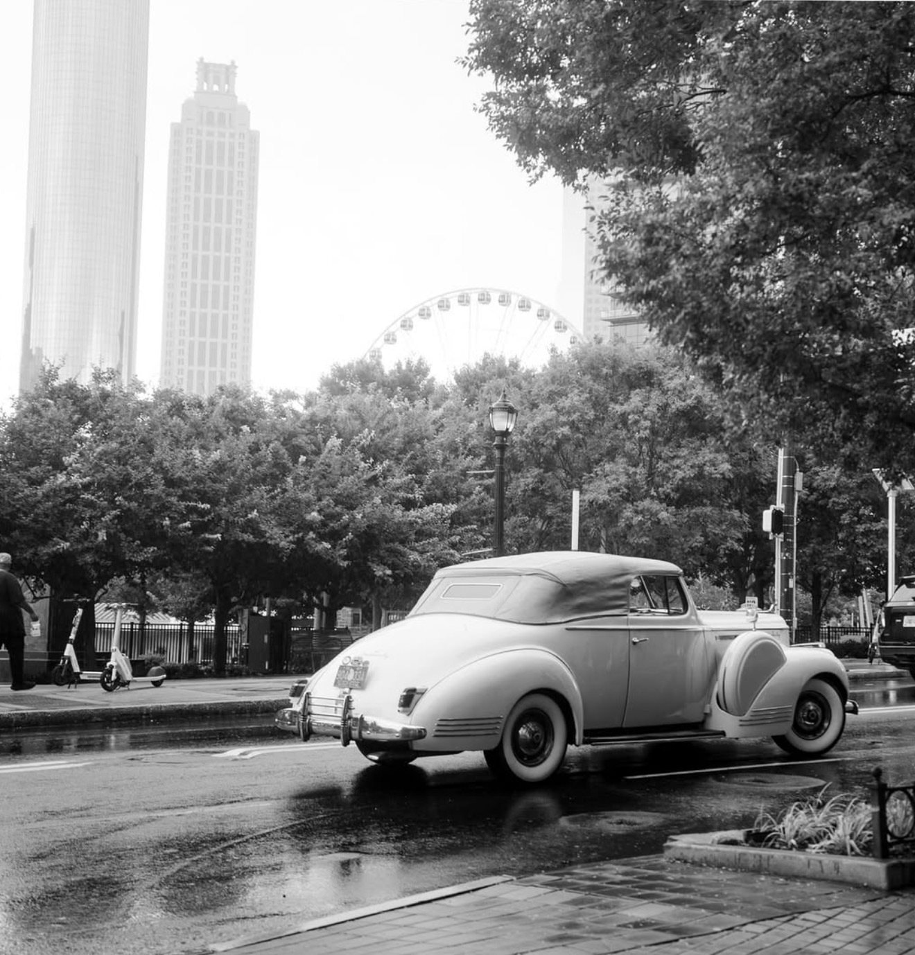 A black-and-white photo of a vintage convertible parked on a city street, with tall buildings and a Ferris wheel behind it.