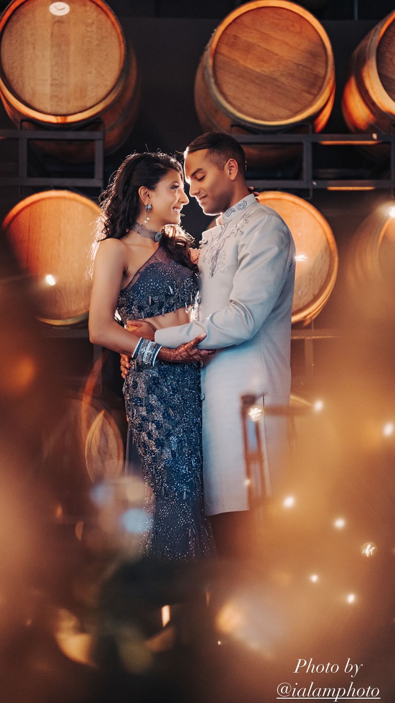 A couple embraces in a wine cellar lined with oak barrels, the woman in a beaded slate-blue gown and the man in a pale grey sherwani, with warm bokeh lights in the foreground.